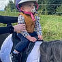 child, baby, helmet, pacifier, horse, saddle, outdoor, grass, trees, person, hand, plaid_shirt, vest, shoes, nature, animal, riding, pony, braided_hair, sky