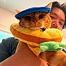 Teri is registered to the contest to win money with this photo: cat, orange_cat, pet, animal, feline, blue_hat, costume, plush_toy, taco_float, green_plush, human, face, whiskers, eyes, indoors, portrait, close_up, funny, playful, selfie