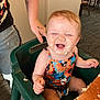 baby, child, high_chair, smiling, happy, face_paint, frosting, food, messy, tattoo, arm, table, chair, indoor, carpet, person, colorful_clothing, dining_room, laughter, cute