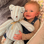 Kolton joined the competition — help win amazing prizes! baby, child, teddy_bear, toy, smile, happy, face, blue_eyes, denim_overalls, sweater, cushion, chair, indoor, plush, patterned_fabric, person, infant, cute, cozy, comfort