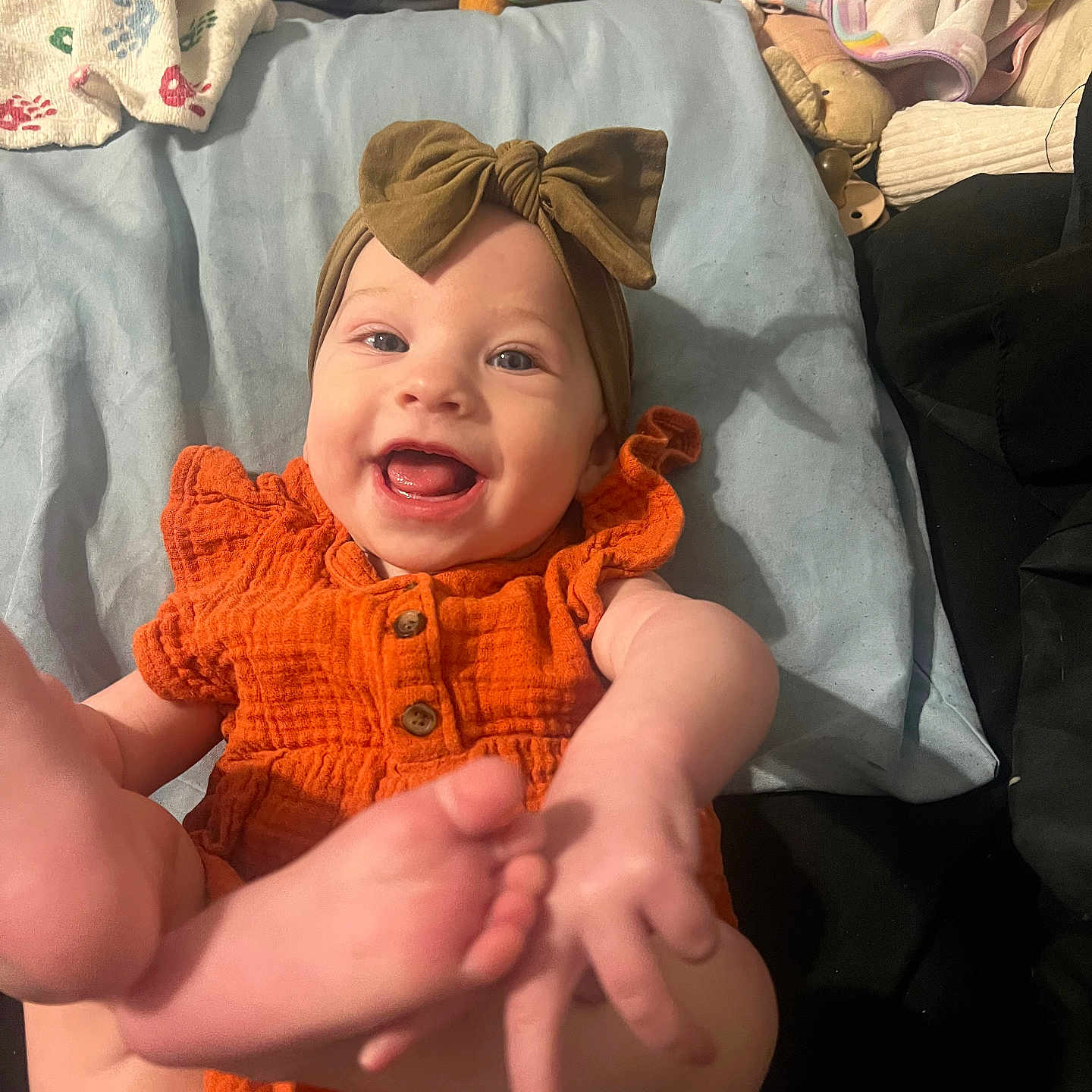 Nova is registered to the contest to win money with this photo: baby, bed, blanket, child, cozy, cute, feet, hand, happy, headband, indoor, infant, kicking_feet, lying_down, orange_outfit, person, playful, portrait, smiling, stuffed_toy