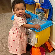 Samara is registered to the contest to win money with this photo: toddler, child, pink_dress, cherry_print, toy_kitchen, play, indoor, carpet, smile, happy, barefoot, furniture, colorful, plastic_toy, curly_hair, person, young_child, home, cute, playtime
