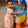 toddler, child, pink_dress, cherry_print, toy_kitchen, play, indoor, carpet, smile, happy, barefoot, furniture, colorful, plastic_toy, curly_hair, person, young_child, home, cute, playtime