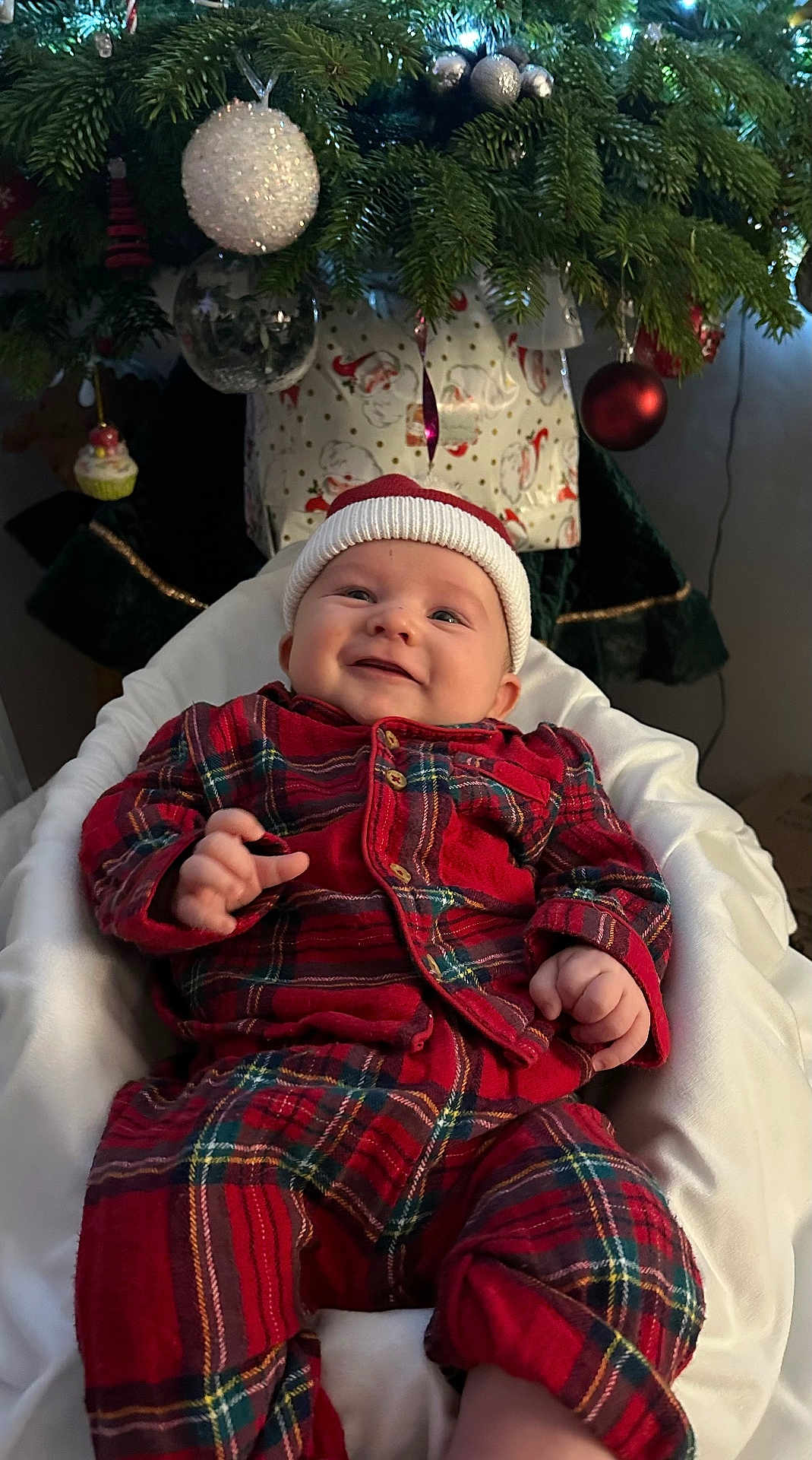 Levi joined the competition — help win amazing prizes! baby, infant, smiling, red_pajamas, santa_hat, christmas_tree, ornaments, holiday, festive, decorations, lights, indoor, white_cushion, cozy, cute, child, seasonal, happy, portrait, celebration