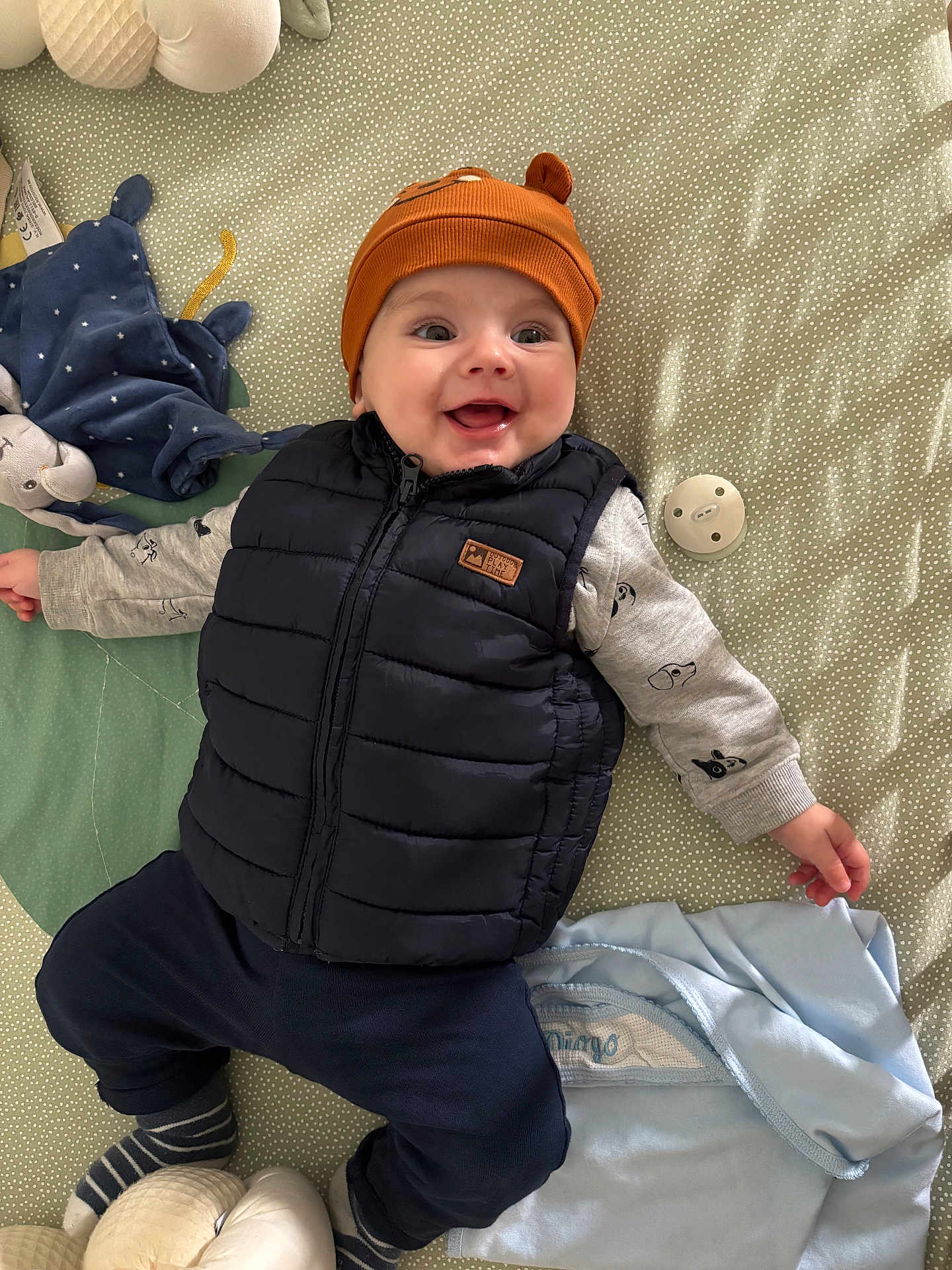 Diogo a rejoint le concours — aidez-le/la à gagner de superbes lots ! baby, blanket, child, clothing, cute, face, happy, hat, headwear, indoor, infant, lying_down, pacifier, patterned_blanket, puffer_vest, smiling, soft_toys, striped_socks, toys, warm