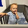 toddler, child, crib, baby, onesie, curly_hair, blue_bedding, sea_animals, bedroom, indoor, furniture, person, hand, face, sucking_fingers, cute, standing, gray_crib, white_wall, blue_tape