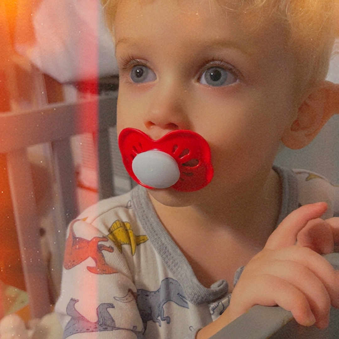 Liam is registered to the contest to win money with this photo: toddler, child, pacifier, crib, pajamas, dinosaur_print, blond_hair, blue_eyes, indoor, baby, portrait, hand, sleepwear, bedroom, soft_lighting, closeup, cute, infant, expression, home