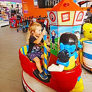 Riley joined the competition — help win amazing prizes! child, toddler, arcade_ride, indoor, shopping_mall, colorful, toy, play_area, smiling, happy, blonde_hair, shorts, sneakers, seat, pole, strollers, retail_shelves, floor_tiles, bear, amusement_ride