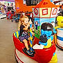 child, toddler, arcade_ride, indoor, shopping_mall, colorful, toy, play_area, smiling, happy, blonde_hair, shorts, sneakers, seat, pole, strollers, retail_shelves, floor_tiles, bear, amusement_ride
