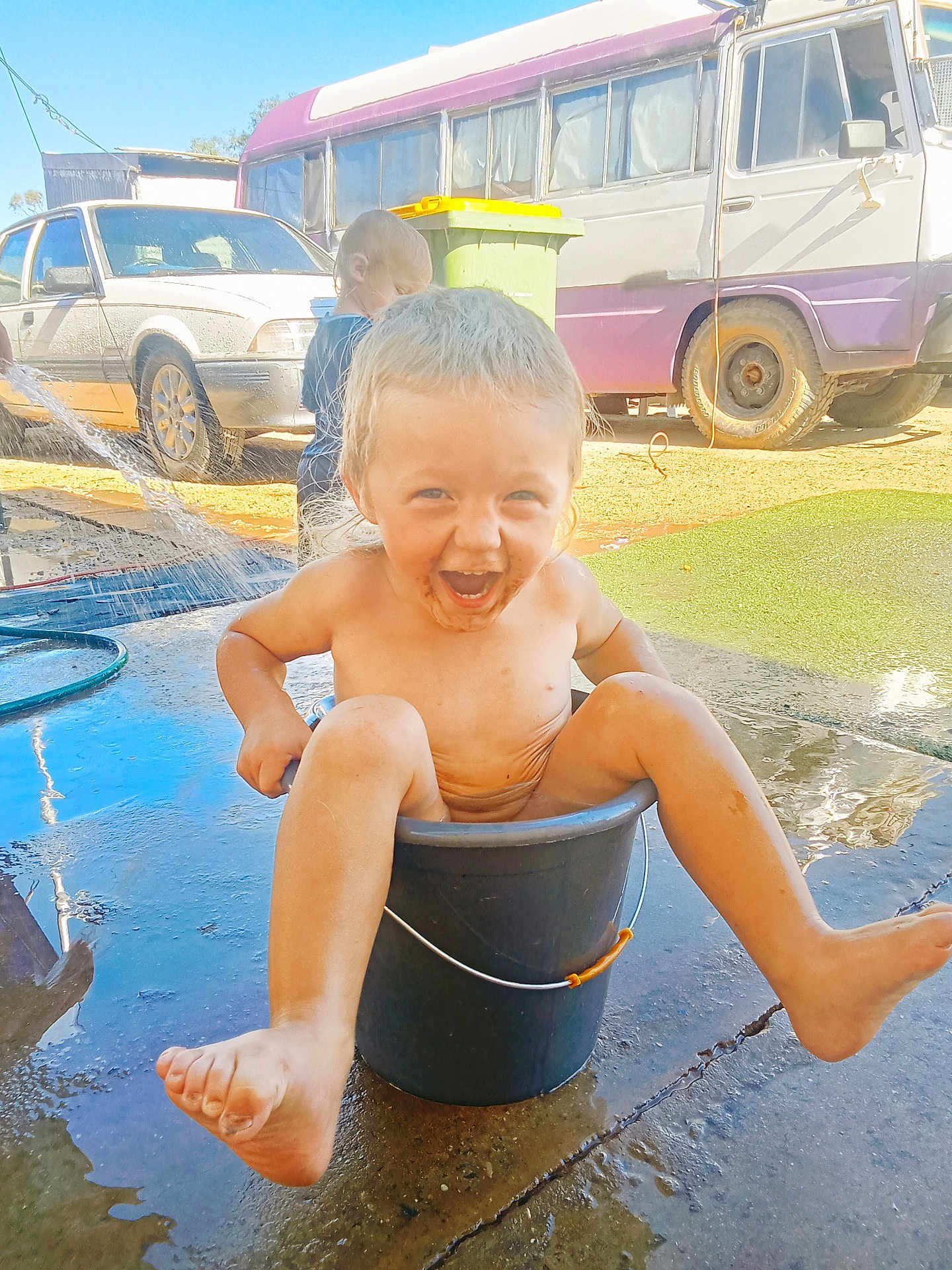 Riley is registered to the contest to win money with this photo: barefoot, bucket, car, child, concrete, happy, hose, laughing, outdoor, playing, playtime, portrait, smiling, sunlight, toddler, trash_bin, van, vehicle, water, wet