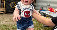 Ethan Mateo is registered to the contest to win money with this photo: baby, child, smiling, outdoor, grass, hat, blue_cap, shorts, shoes, person, hand, adult, picnic_table, toys, building, wooden_structure, casual_clothing, summer, happy, playful