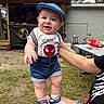adult, baby, blue_cap, building, casual_clothing, child, grass, hand, happy, hat, outdoor, person, picnic_table, playful, shoes, shorts, smiling, summer, toys, wooden_structure