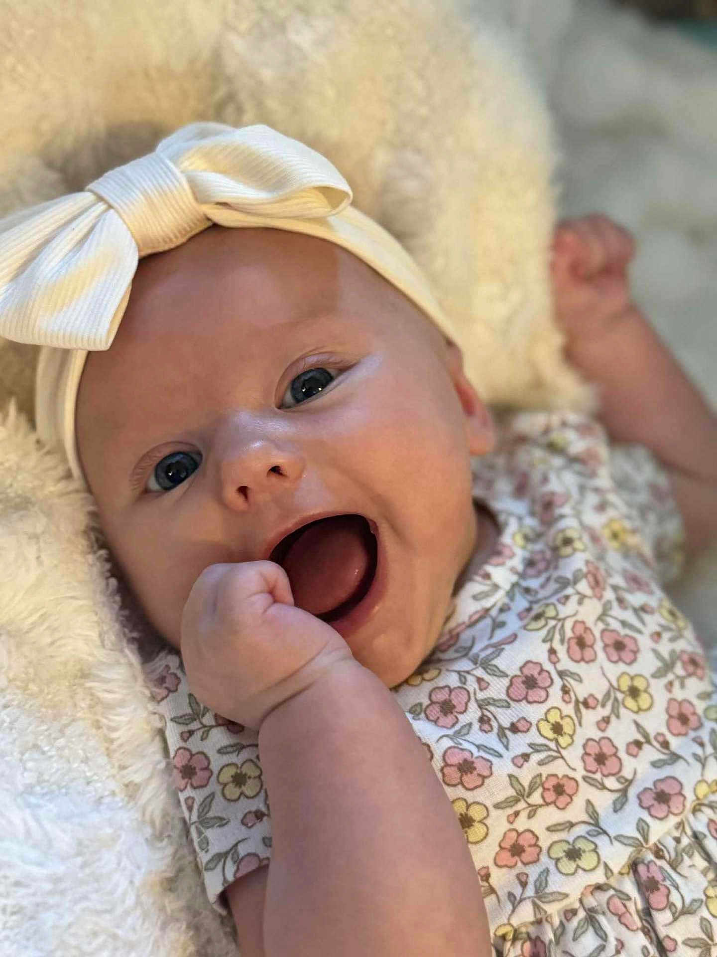 Lily is registered to the contest to win money with this photo: baby, infant, smiling, headband, bow, floral_dress, hand, face, blue_eyes, skin, blanket, soft_texture, cute, happy, portrait, closeup, child, newborn, expression, lying_down