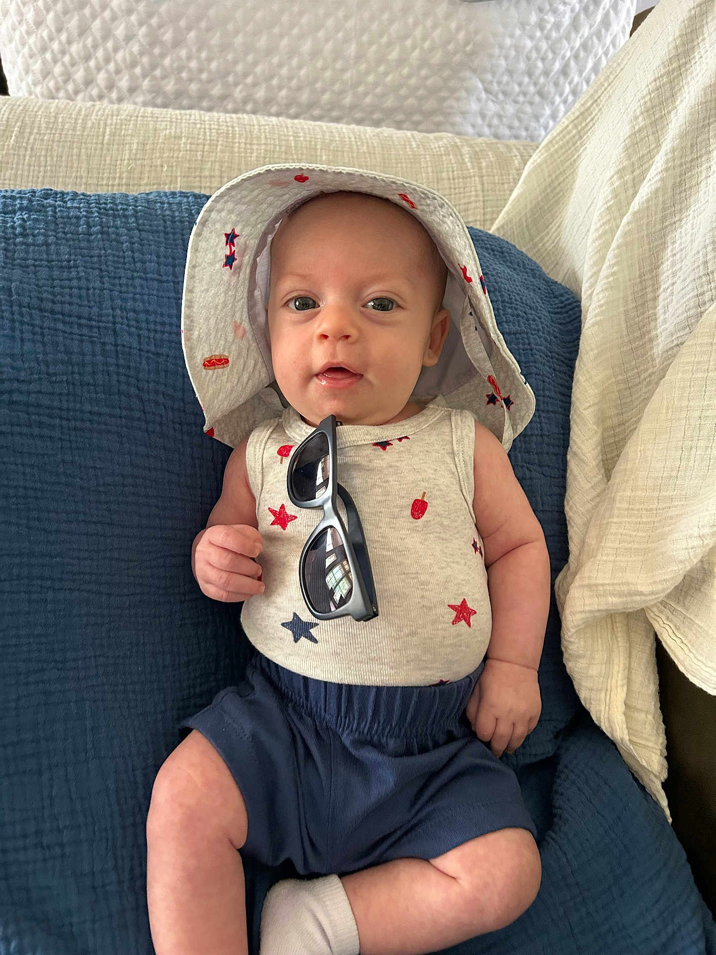 Rj joined the competition — help win amazing prizes! baby, infant, hat, sunglasses, tank_top, shorts, cushion, quilt, indoor, clothing, face, person, smile, cute, relaxing, seated, skin, headwear, footwear, comfort