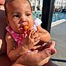 baby, child, infant, portrait, snack, carrot, eating, pink_clothing, bib, polka_dots, adult_hands, holding, poolside, swimming_pool, outdoor, sunlight, water, buildings, close_up, happy