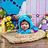 Noa is registered to the contest to win money with this photo: baby, infant, child, monster_costume, blue, hood, smiling, face, portrait, blanket, knitted_blanket, toys, stuffed_animals, playroom, indoor, colorful, sitting, cute, costume, photography