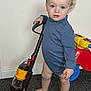 toddler, child, blue_onesie, toy_vacuum, carpet, indoor, barefoot, ride_on_toy, curly_hair, standing, playtime, young_child, floor, wall, hand, face, expression, toy, person, home