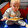 Alfie is registered to the contest to win money with this photo: toddler, child, book, reading, curious, indoor, sitting, nike_sweatshirt, blonde_hair, expression, padded_surface, play_area, clothing, casual, young, person, face, hand, open_mouth, engaged