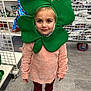 child, girl, face, green_shamrock_mask, costume, store, aisle, shelf, snacks, clothing, pink_sweater, maroon_pants, shoes, floor, shopping_cart, smile, portrait, indoor, retail, holiday