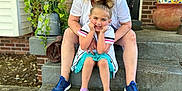 Angel is registered to the contest to win money with this photo: child, woman, smiling, steps, porch, brick_wall, plants, potted_plant, door, curly_hair, casual_clothing, sneakers, purple_shoes, blue_shoes, outdoor, daylight, happy, family, together, portrait
