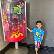 Joshua Castillo is registered to the contest to win money with this photo: advertisement, blue_shirt, boy, casual, child, colorful, crocs, floor, happy, indoor, mario, mcdonalds, plaid_shorts, princess_peach, promotional_display, smile, standing, super_mario_galaxy, tiled_wall, yoshi