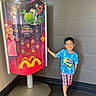 child, boy, smile, blue_shirt, plaid_shorts, crocs, mcdonalds, promotional_display, super_mario_galaxy, princess_peach, yoshi, mario, indoor, tiled_wall, floor, advertisement, colorful, happy, casual, standing