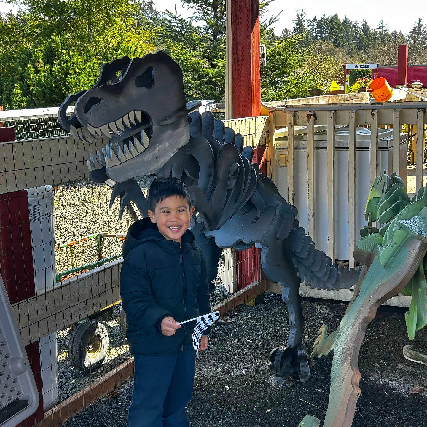 Joshua Castillo is registered to the contest to win money with this photo: child, boy, smile, jacket, sneakers, dinosaur, wooden_sculpture, outdoor, fence, trees, flag, playground, daylight, happy, person, nature, wood, sculpture, toy, recreation