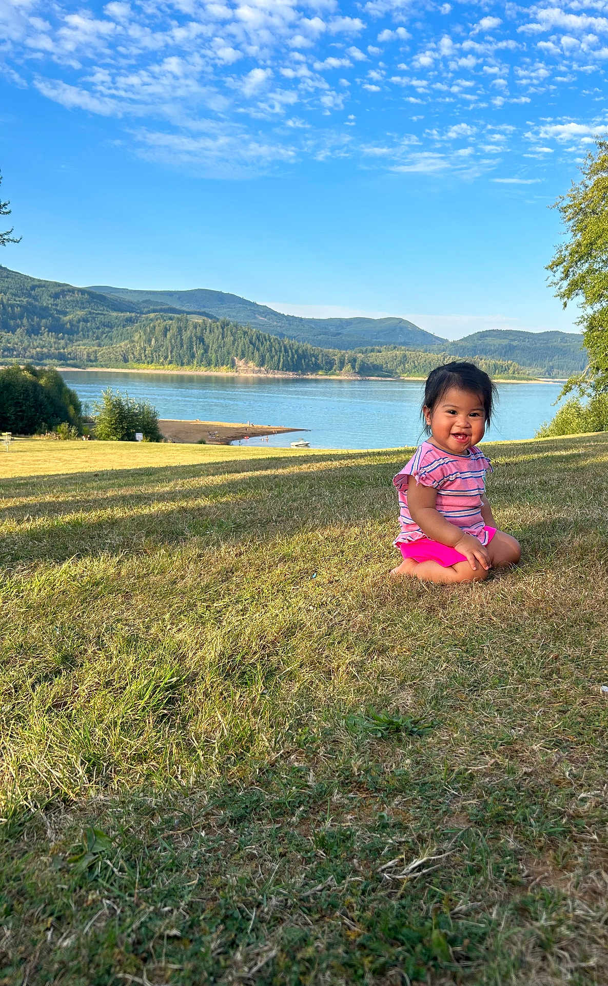 Gabriela is registered to the contest to win money with this photo: toddler, child, grass, lake, water, hill, mountain, trees, sky, clouds, outdoor, nature, smiling, happy, sitting, sunlight, casual_clothing, landscape, summer, daytime