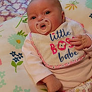 Aiyana is registered to the contest to win money with this photo: baby, pacifier, bib, floral_blanket, infant, cute, lying_down, pink_clothing, hand, face, head, person, indoors, soft_toys, patterned_pants, newborn, child, sleeping_area, colorful, cozy