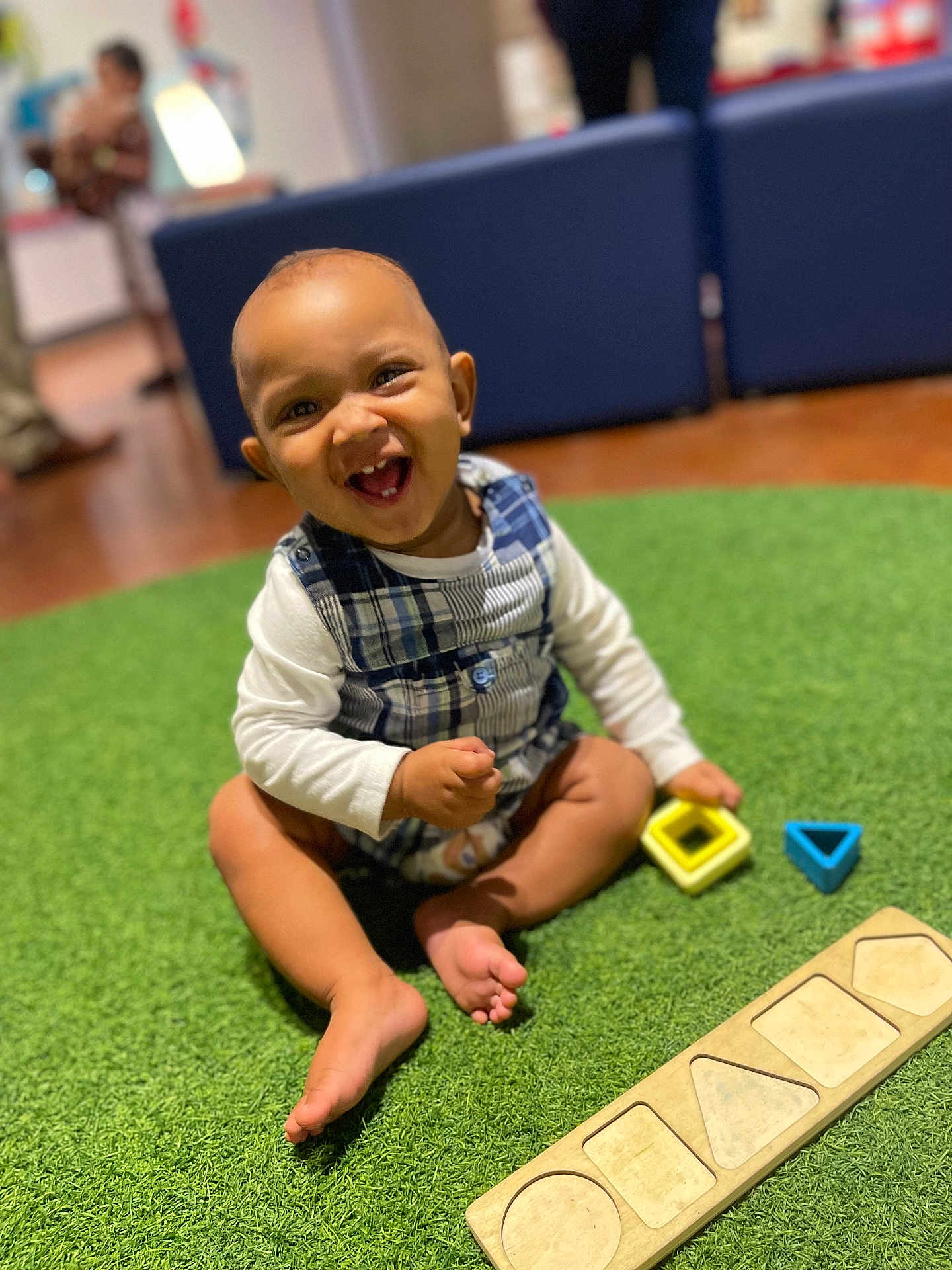 Malachi is registered to the contest to win money with this photo: toddler, child, smiling, playing, toy, shape_blocks, indoor, carpet, green, happy, sitting, baby, clothing, plaid, foot, furniture, blurred_background, person, room, cute