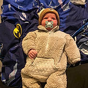 Leo is registered to the contest to win money with this photo: baby, infant, pacifier, onesie, knit_hat, blanket, batman_logo, blue_blanket, soft_clothing, cute, child, indoor, lying_down, face, headwear, comfort, warm_clothing, expression, foot, person