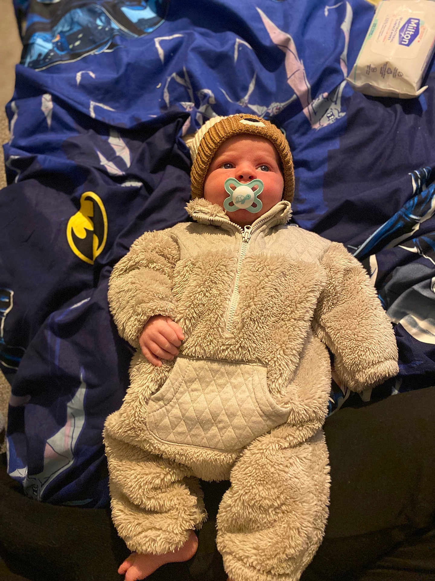 Leo is registered to the contest to win money with this photo: baby, infant, pacifier, onesie, knit_hat, blanket, batman_logo, blue_blanket, soft_clothing, cute, child, indoor, lying_down, face, headwear, comfort, warm_clothing, expression, foot, person