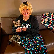 Danni Slay is registered to the contest to win money with this photo: person, cat, tabby_cat, pet, indoors, living_room, couch, leather_sofa, holiday, christmas, festive, pajamas, light_bulbs, gingerbread, blanket, smiling, portrait, sitting, companion, cozy