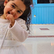 Manmeet joined the competition — help win amazing prizes! child, girl, blurry, white_dress, pink_bow, curly_hair, indoor, blue_door, cafe_bar_sign, floor_tiles, playful, smiling, hand_near_mouth, young_child, person, portrait, casual, cute, decor, lighting