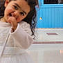 child, girl, blurry, white_dress, pink_bow, curly_hair, indoor, blue_door, cafe_bar_sign, floor_tiles, playful, smiling, hand_near_mouth, young_child, person, portrait, casual, cute, decor, lighting