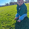 Lis participe au concours pour gagner de l'argent avec cette photo : baby, child, grass, outdoor, sunlight, shadow, green_jacket, barefoot, blue_pants, field, daylight, sky, trees, buildings, nature, casual_clothing, sitting, portrait, young_child, curious