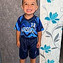 background, blue, boy, casual, child, flooring, floral_pattern, happy, home, indoor, jersey, person, portrait, shorts, smiling, sneakers, sportswear, standing, wallpaper, young