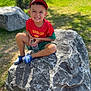 boy, cap, casual_clothing, child, daytime, grass, greenery, happy, nature, outdoor, park, person, playful, red_shirt, rock, shoes, shorts, smiling, sunlight, water_bottle