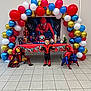 Yanis a rejoint le concours — aidez-le/la à gagner de superbes lots ! child, spiderman, costume, balloons, party, decorations, toys, action_figures, superhero, red, blue, gold, white, floor, tiles, table, banner, candy, celebration, indoor