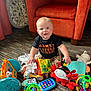 baby, child, toys, colorful_toys, carpet, orange_armchair, indoor, smiling, sitting, black_shirt, patterned_pants, plush_toy, rattle, floor, happy, face, hand, foot, room, playtime