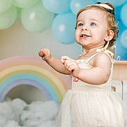 Hadleigh is registered to the contest to win money with this photo: toddler, child, blue_eyes, smiling, white_dress, bow, balloons, rainbow, clouds, pastel_colors, portrait, indoor, celebration, happy, cute, person, baby, curly_hair, background, festive