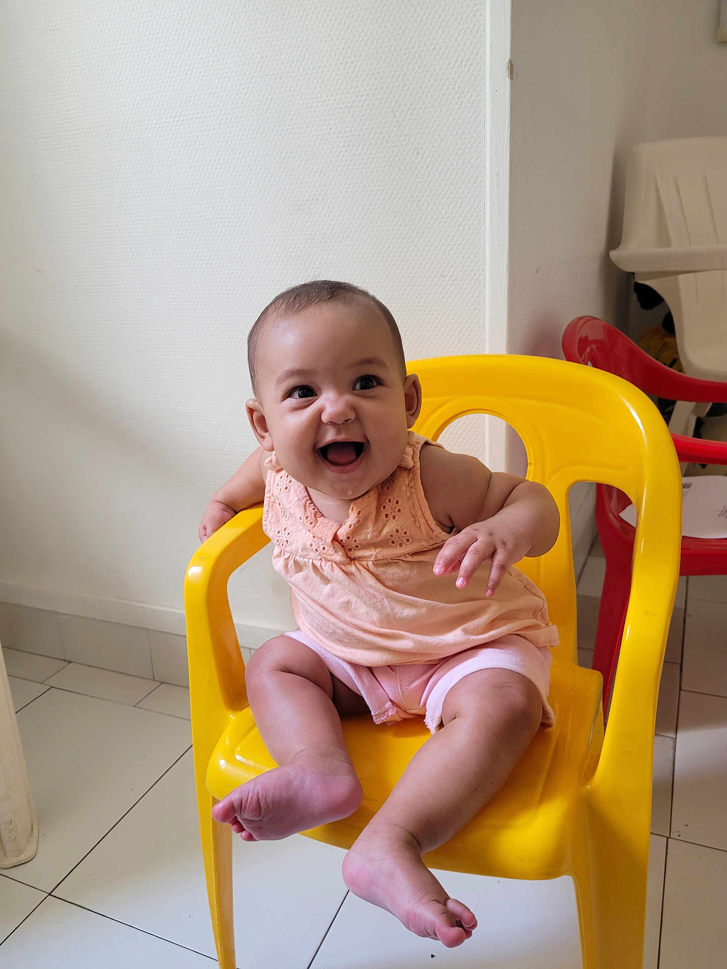 Athéna a rejoint le concours — aidez-le/la à gagner de superbes lots ! baby, child, smiling, yellow_chair, plastic_chair, indoor, floor_tiles, casual_clothing, happy, cute, seated, infant, person, shorts, top, footwear_free, armrest, head, face, playful