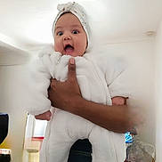 Athéna participe au concours pour gagner de l'argent avec cette photo : baby, child, indoor, person, white_clothing, headband, hand, surprised, expression, fluffy_outfit, wide_eyes, open_mouth, holding, adult_hand, cozy, room, casual, portrait, cute, infant
