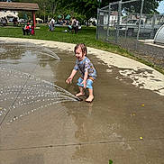 Kylo is registered to the contest to win money with this photo: child, water, splash_pad, playing, barefoot, wet, summer, park, grass, trees, playground, people, outdoor, fun, joyful, clothing, concrete, fence, water_spray, daytime