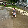 child, water, splash_pad, playing, barefoot, wet, summer, park, grass, trees, playground, people, outdoor, fun, joyful, clothing, concrete, fence, water_spray, daytime