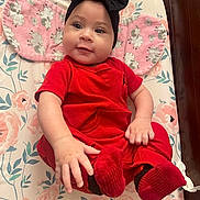 Avory is registered to the contest to win money with this photo: baby, child, red_clothing, black_headband, socks, floral_pattern, changing_pad, cute, infant, smiling, indoors, person, baby_girl, headwear, hand, footwear, fabric, soft_texture, wooden_furniture, comfort