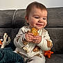 baby, child, smile, couch, blanket, toy, onesie, food, bread, happy, indoor, infant, cute, sitting, face, hand, cozy, expression, playful, home