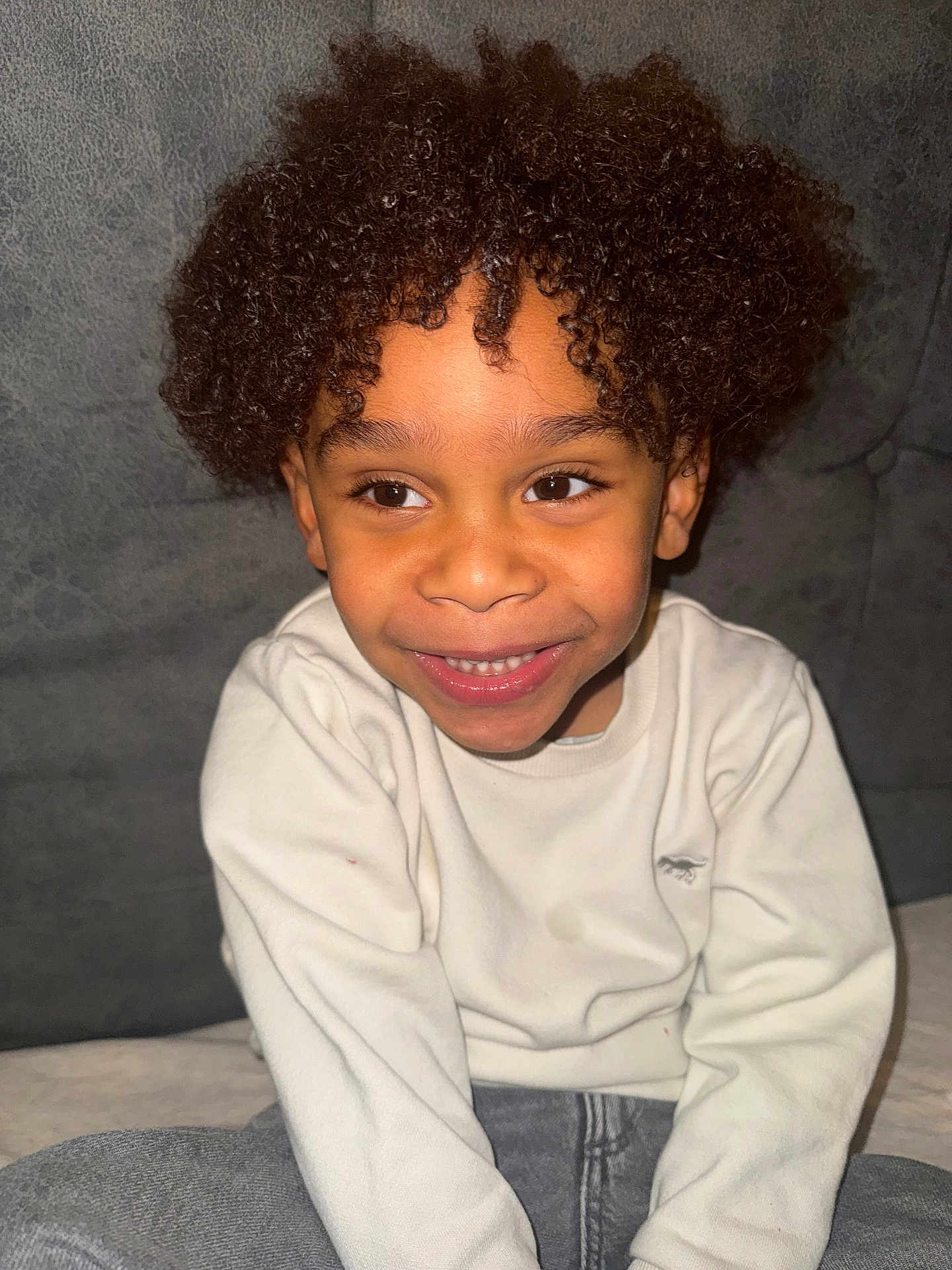 Kingsley participe au concours pour gagner de l'argent avec cette photo : child, smiling, curly_hair, sitting, white_sweatshirt, gray_pants, portrait, happy, face, young, indoors, casual_clothing, curly_afro, cute, person, joy, comfort, closeup, childhood, warmth