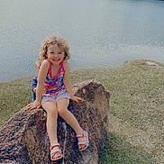 Elizabeth is registered to the contest to win money with this photo: child, smiling, rock, lake, water, fountain, grass, outdoor, nature, trees, curly_hair, sandals, summer, happy, playful, casual_clothing, daytime, scenic, relaxing, person