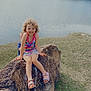 child, smiling, rock, lake, water, fountain, grass, outdoor, nature, trees, curly_hair, sandals, summer, happy, playful, casual_clothing, daytime, scenic, relaxing, person